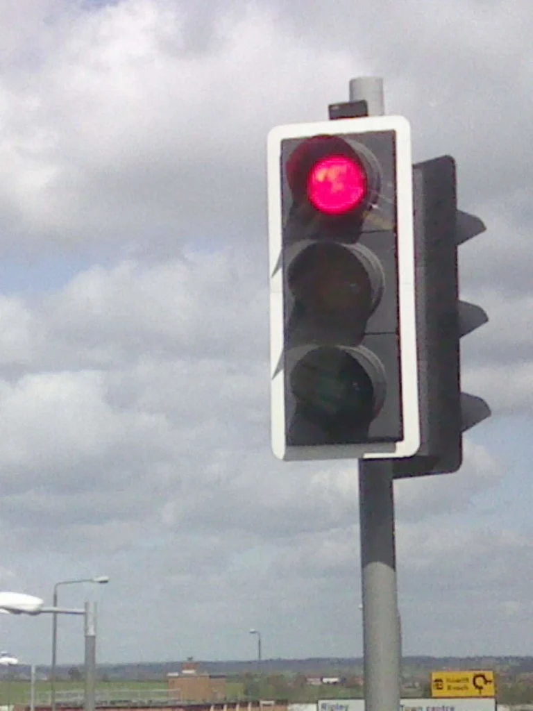 A British traffic light on a grey pole, mounted on a black backboard with a white border, shows only its red lamp lit against a cloudy sky, with a street lamp and a small yellow road sign low in the frame.