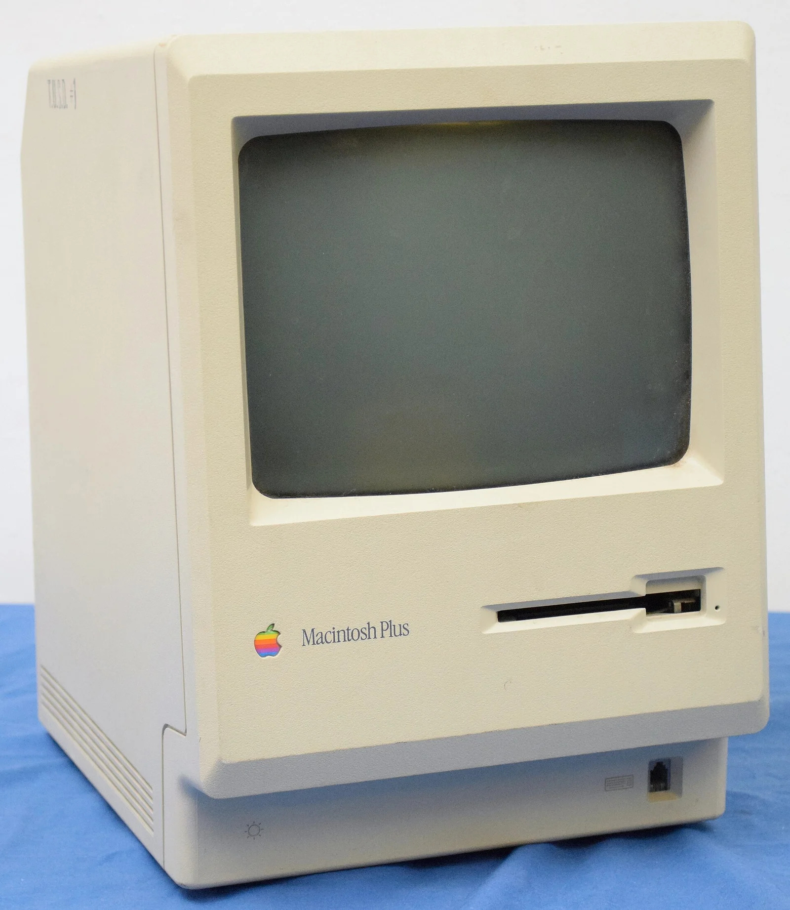 A beige Apple Macintosh Plus computer with a dark screen stands on a blue cloth.