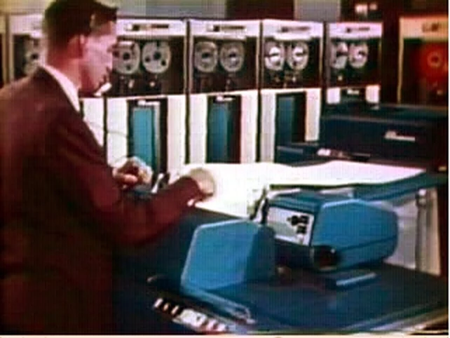 A black and white photograph of a row of tall reel-to-reel tape drives in a 1960s machine room.